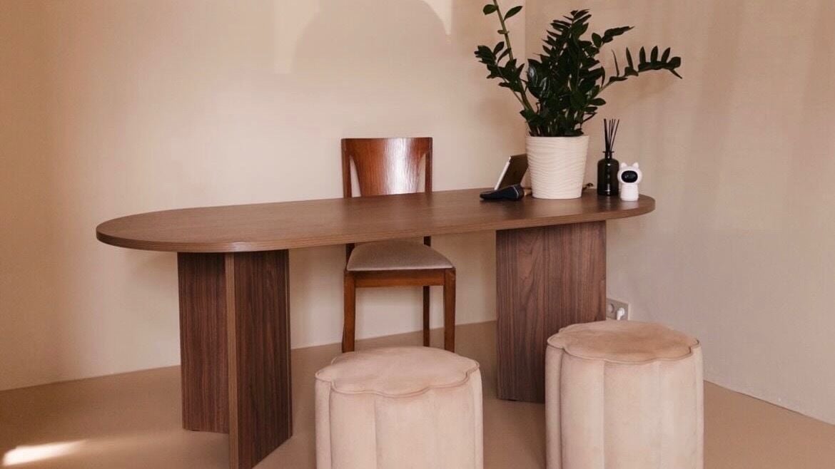Het consulthoekje: een ovale houten tafel, een stoel, twee ronde poefs en een plant tegen een zachtroze muur